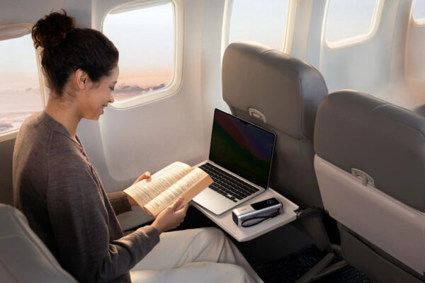 Anker power bank on aeroplane tray table with laptop and person reading a book