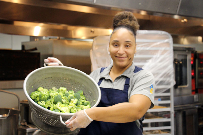 A woman smiles as she shows a very large colander filled with broccoli. She is wearing a gray shirt and blue apron.