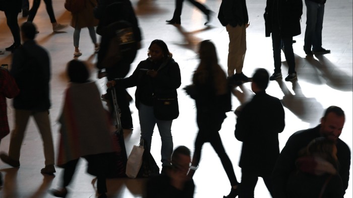 Silhouettes of people walking and standing in Grand Central Terminal during morning rush hour, some carrying bags or rolling luggage.