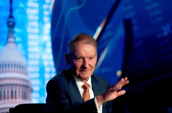 Paul Atkins gestures while seated, with a backdrop featuring the US Capitol dome and financial graphics.