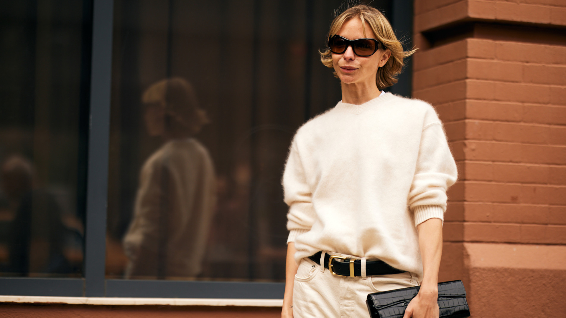 Fashion week spring 2026 attendee wearing sunglasses, white v-neck sweater, black belt, white pants, black clutch