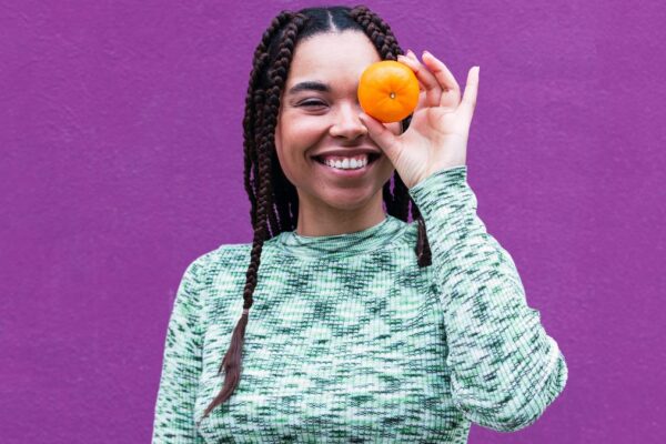 A person with long brown braided hair and a teal patterned shirt holding an orange up in front of their left eye.