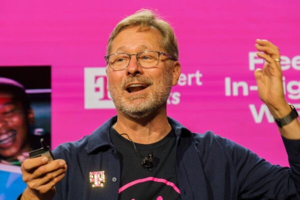 A bearded man speaks on stage with one arm raised and a remote control in the other hand.