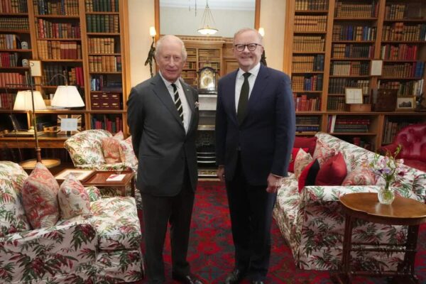 Two men stand in a richly appointed library, flanked by tall wooden bookshelves filled and armchairs and sofas. The man on the left (King Charles) wears a grey suit and striped tie, while the man on the right (Anthony Albanese) wears a dark blue suit and a solid dark tie, both posing directly for the camera.