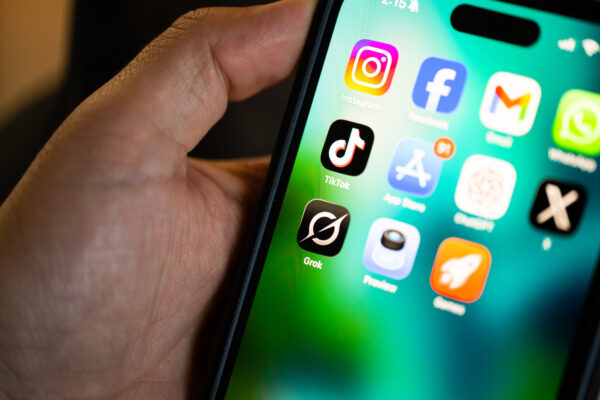 Could Elon Musk’s Grokipedia Mean Trouble for Wikipedia? A hand holding an iPhone showing several apps on the home screen.