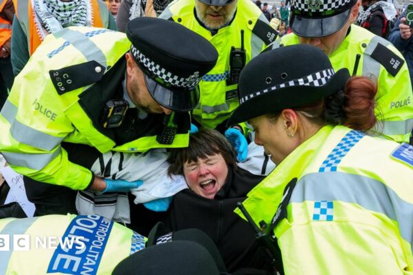 Three male police officers detain a female protester, who is holding a paper sign appearing to mention Palestine, during a rally challenging the UK government's proscription of "Palestine Action" under anti-terrorism law. In the background there are grand buildings a Palestine flag and a poster mentioning genocide