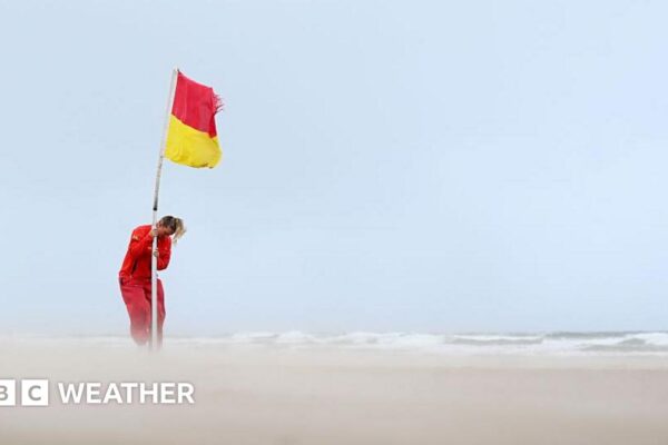 Lifeguard securing a red and yellow flag on a deserted beach with sand whipped up by strong winds