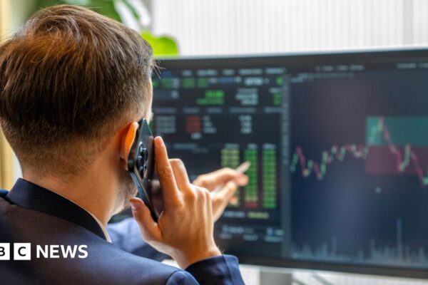 A man with his back to the camera holds a mobile phone to his ear and points to computer screens showing financial market information