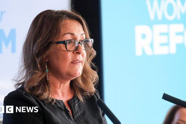 Linden Kemkaran wearing glasses and talking into two podium microphones. Light blue and white backgrounds can be seen behind her.