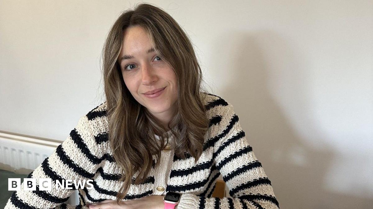 Abbi, a young woman with dark blonde hair. She is wearing a black and white striped cardigan and is sitting at her kitchen table