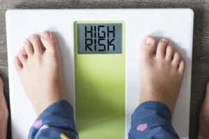 A photo of a child's feet on a weight scale with a woman's feet behind them and the words "high risk" on the scale display.