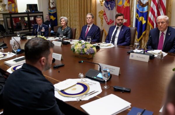 Volodymyr Zelenskyy and Donald Trump sit across from each other at a formal meeting table with officials during a White House bilateral meeting.