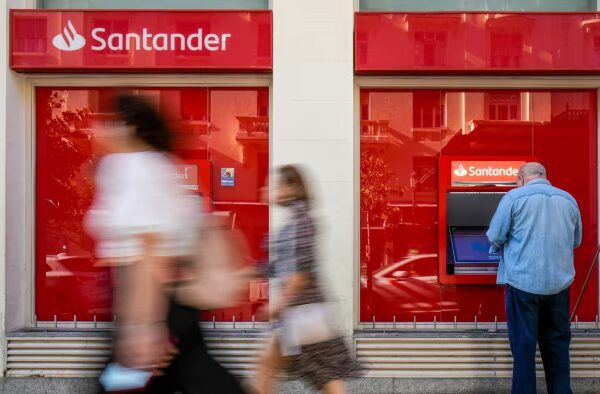 A man uses an ATM outside a Banco Santander branch in Madrid as several people walk past the bank’s red storefront.
