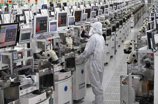 A technician in protective clothing operates equipment among rows of chip manufacturing machines at a semiconductor plant.
