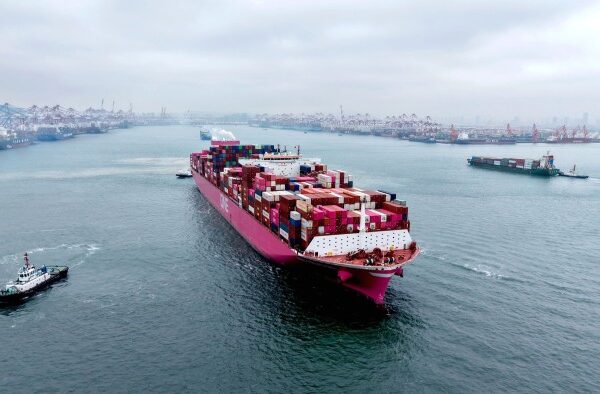A large pink container ship loaded with multicolored shipping containers sails out of a busy port, with cranes and other ships in the background.
