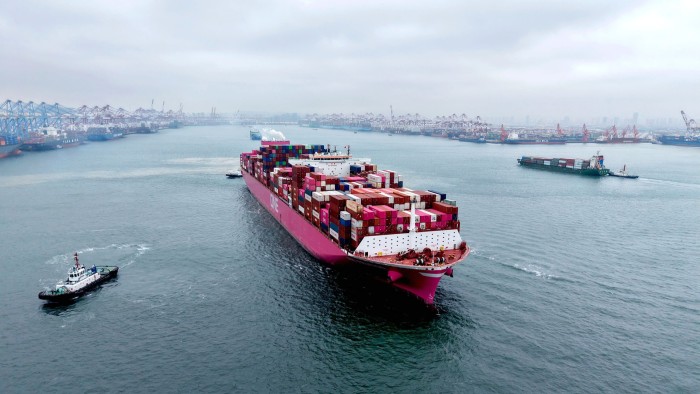 A large pink container ship loaded with multicolored shipping containers sails out of a busy port, with cranes and other ships in the background.