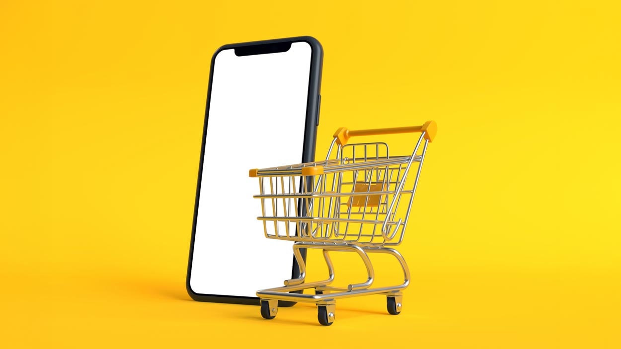 A small shopping cart stands in front of a smartphone with a blank white screen on a bright yellow background, symbolizing online shopping or the convenience of shopping apps.