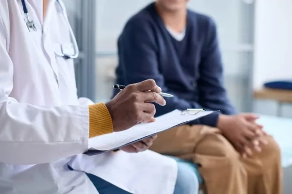 Doctor in a white coat taking notes on a clipboard while sitting with a patient in casual attire