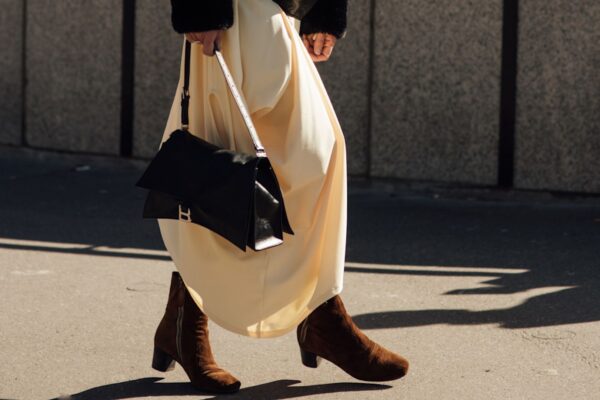 a woman walking on a sidewalk wearing a black fur sleeve jacket, white skirt, brown suede booties, and a black handbag.