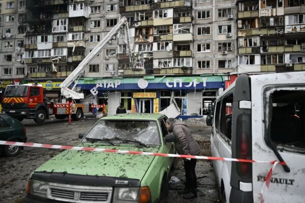A person leans into a green car with a shattered windshield, behind red and white caution tape. A red fire truck extends its ladder toward a multistory building with broken windows.