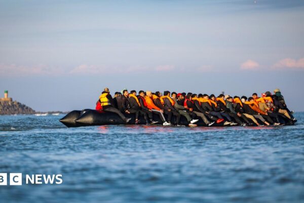 Migrants wearing orange lifevests sit on an inflatable boat on the English Channel, a rocky headland appears in the backdrop
