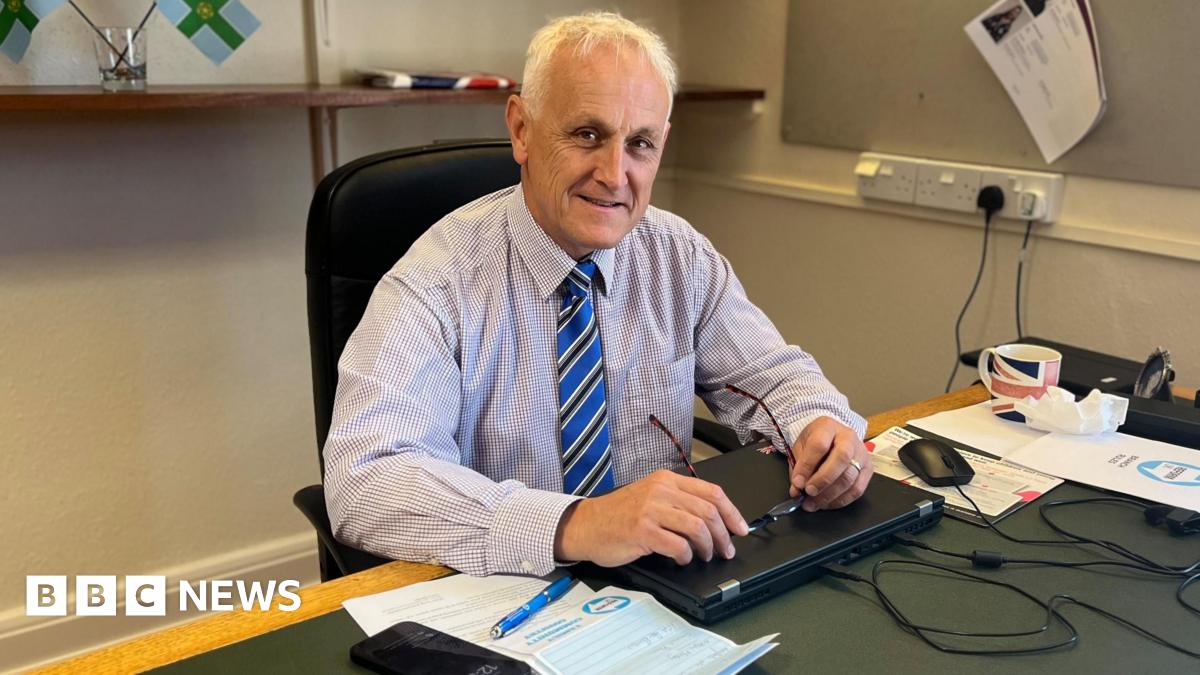Alan Graves (white hair, holding glasses in hands) sits at green topped desk with papers around him. A pair of Derbyshire county flags are on the shelf to his top right.