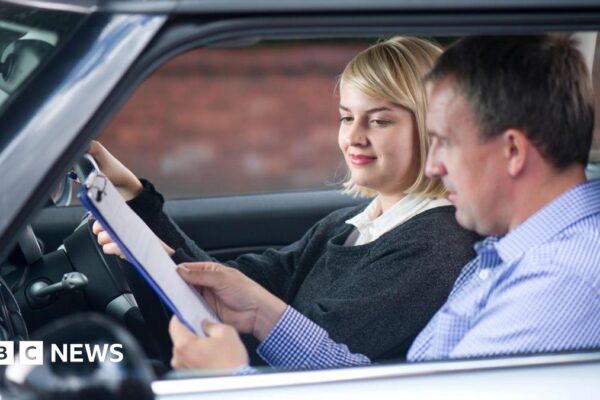 A young woman taking a driving test. She has blonde hair and is wearing a dark grey jumper. Her tester is a man in a check blue shirt. He is showing her a document on a clipboard.
