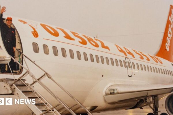 Stelios stands at the top of the plane's steps waving at the camera. He wears an orange Easyjet baseball cap. The side of the plane has the Luton Airport phone number emblazoned in orange against a white plane. Is says 01582 445566. The tail of the plane is orange and has easyjet written in white.