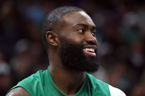 Jaylen Brown smiles, looking forward on the court, wearing a green jersey.