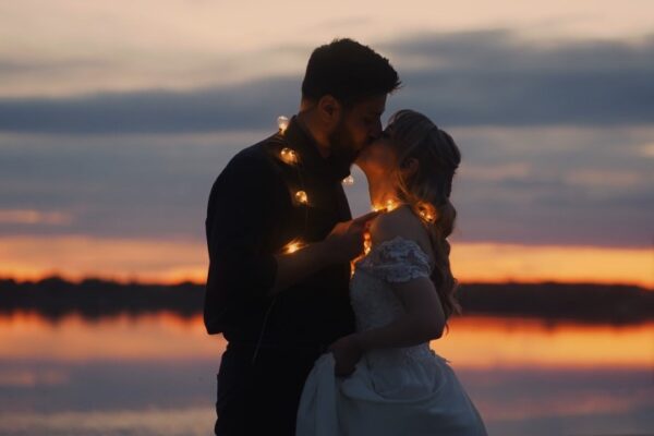A bride and groom share a kiss at sunset, surrounded by twinkling lights creating a romantic atmosphere