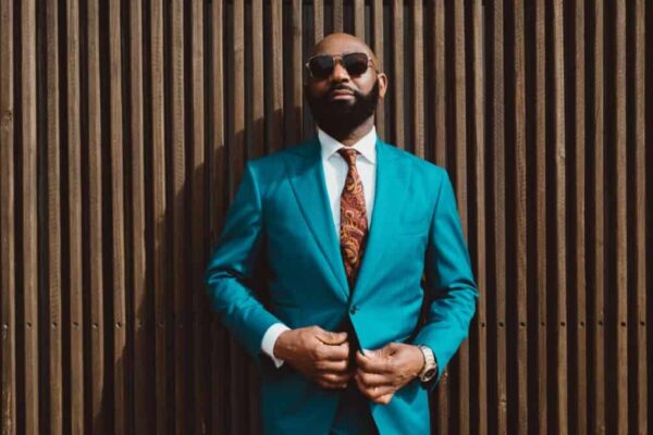 A handsome mature bald bearded black man in a sunglasses and a fashionable blue or teal costume with a tie is standing in front of a wall made of striped wooden timbers and fastening a suit button