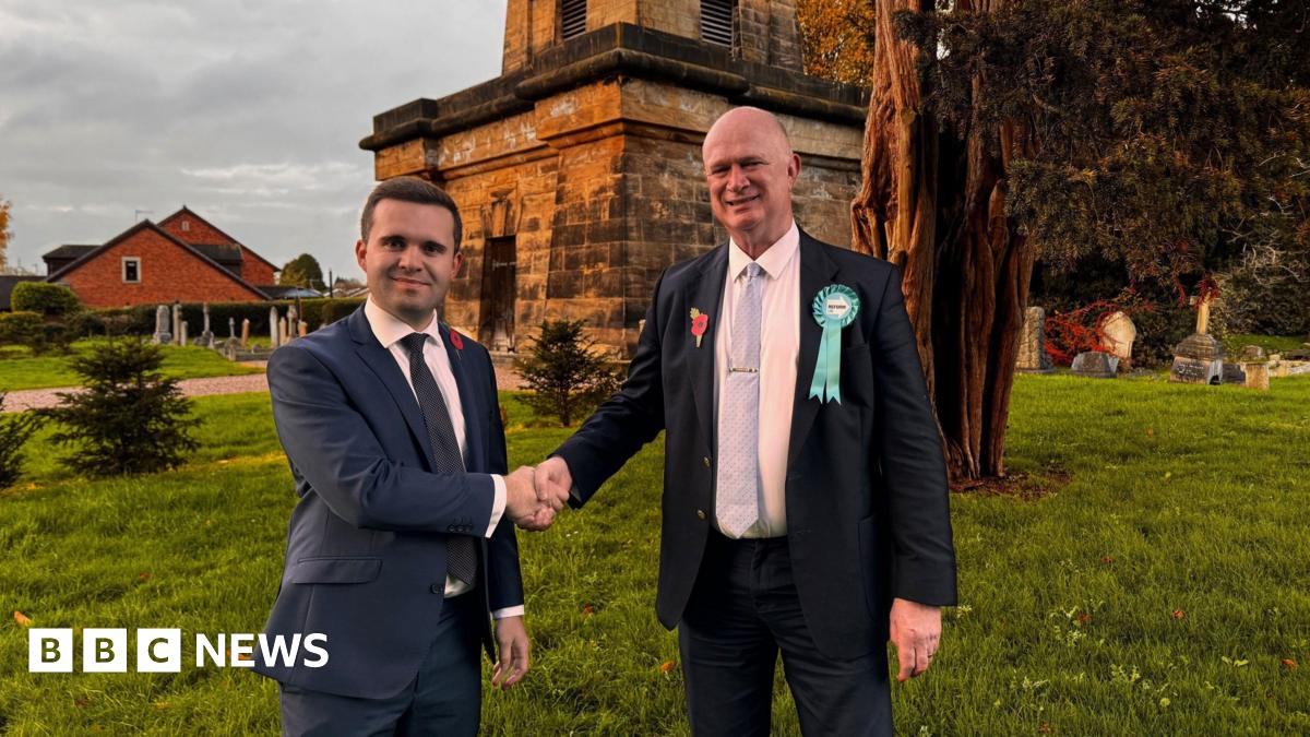 Two men wearing dark suits are seen shaking hands. They are stood in what appears to be a churchyard.