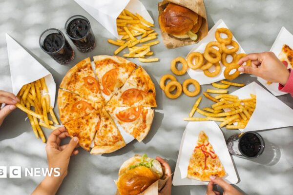 Looking down on a table full of plates of pizza, burgers in buns, onion rings, french fries and cola drinks, which are all examples of ultra-processed foods. Hands are reaching out and picking up the different foods.