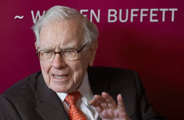 Warren Buffett gestures while speaking, seated in front of a maroon backdrop with his name partially visible.