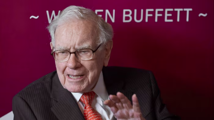 Warren Buffett gestures while speaking, seated in front of a maroon backdrop with his name partially visible.