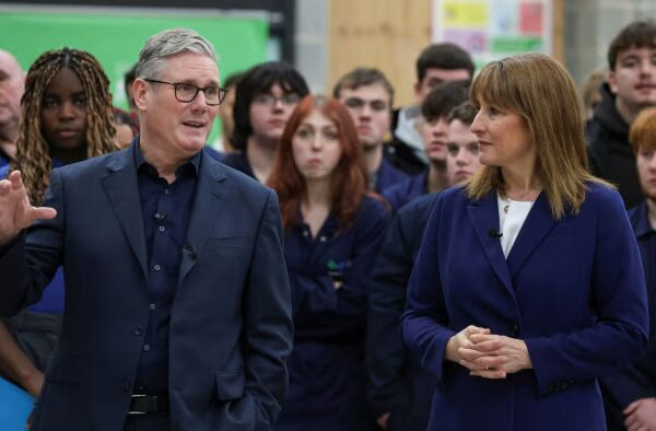 Sir Keir Starmer gestures while speaking beside Rachel Reeves, with a group of students and staff watching in the background