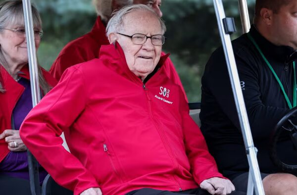 Warren Buffett sits in a golf cart wearing a red jacket with the Allen & Company Sun Valley Conference logo.