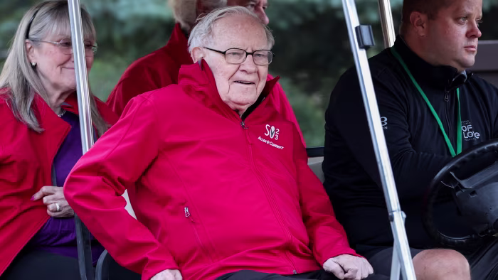 Warren Buffett sits in a golf cart wearing a red jacket with the Allen & Company Sun Valley Conference logo.