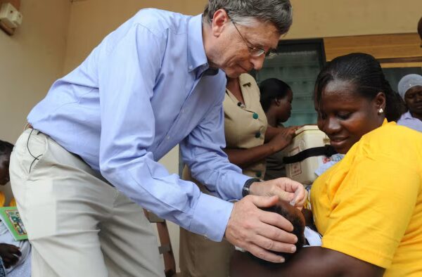 Bill Gates administers a rotavirus vaccine to a baby held by a smiling woman at a health centre in Ghana