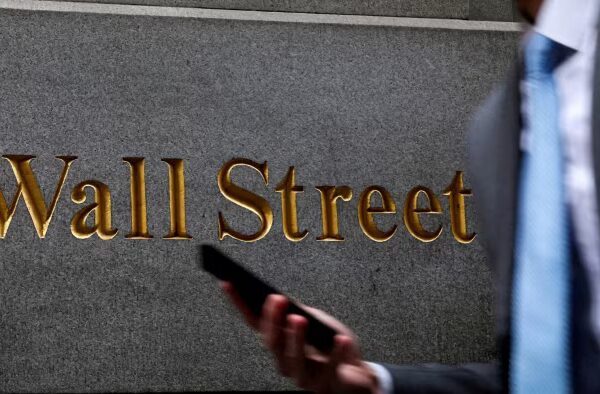 Gold "Wall Street" lettering on a stone wall, with a blurred man in a suit and blue tie holding a phone in the foreground.