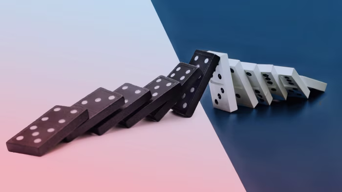 Black and white dominoes falling in a row on a surface divided between pink and blue backgrounds.