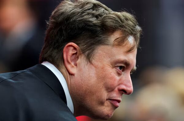 Elon Musk in profile wearing a dark suit, attending an event with a blurred crowd in the background.