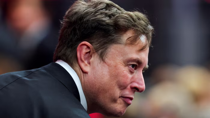 Elon Musk in profile wearing a dark suit, attending an event with a blurred crowd in the background.