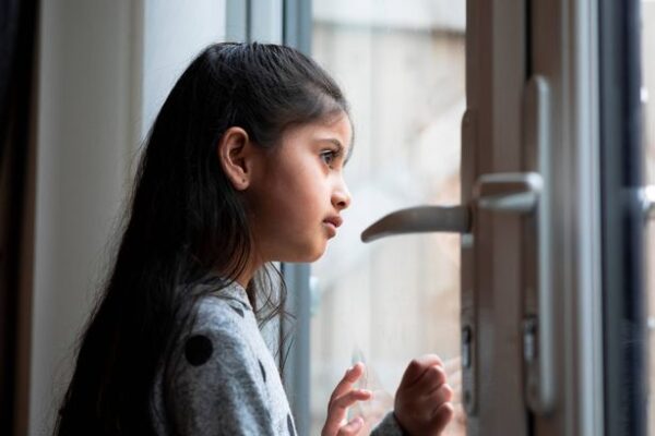A young girl standing at the window and looking out with a blank expression.