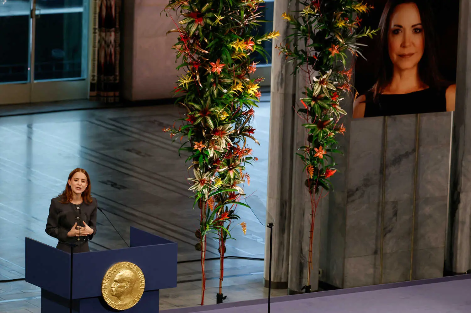 Ana Corina Sosa speaking behind a lectern onstage under a large photo of her mother, María Corina Machado.