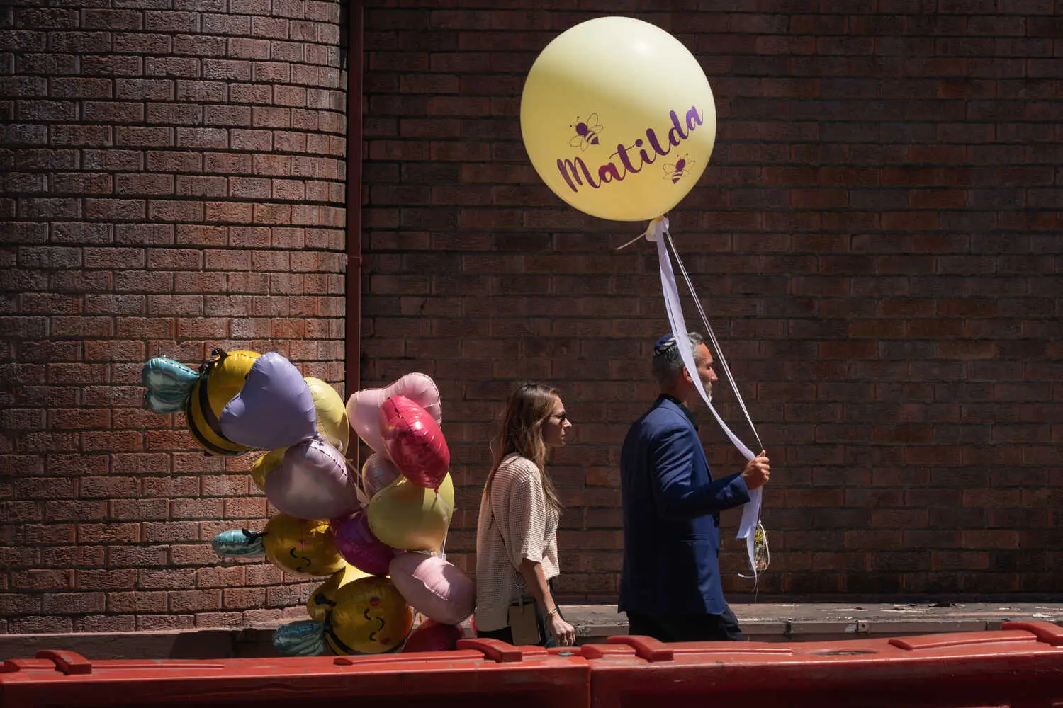 A man carries a large yellow balloon that reads “Matilda,” followed by a woman carrying a bunch of colorful balloons.