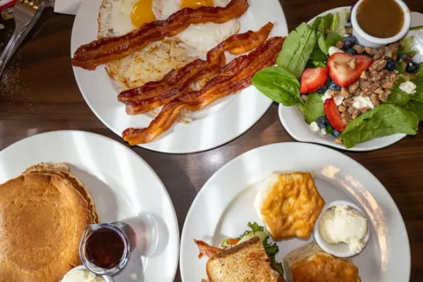 Four plates of diner food; eggs, bacon, pancakes, strawberries, biscuit, toast