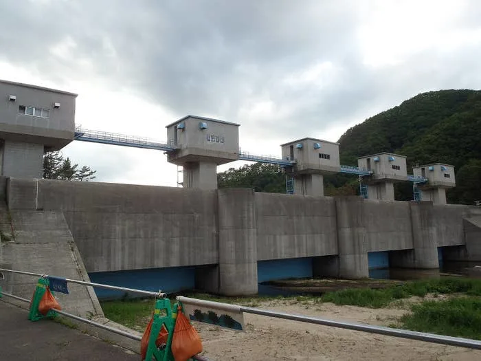 Concrete floodgate structure with four control towers on a river, backed by trees and cloudy sky. Orange sandbags and barriers in foreground
