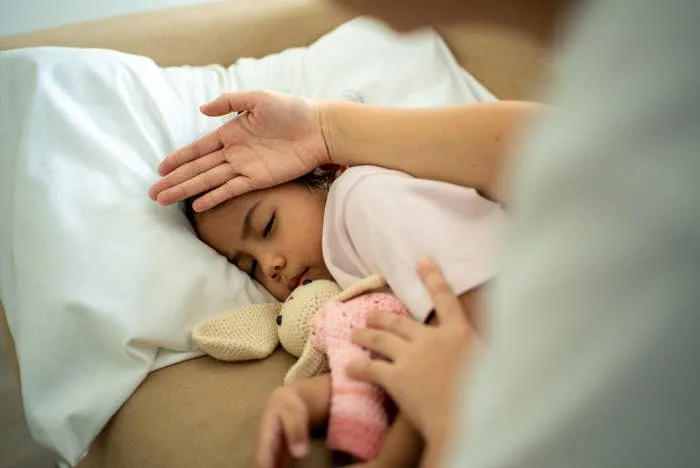 Child sleeping in bed, holding a stuffed toy. An adult hand gently checks the child's forehead