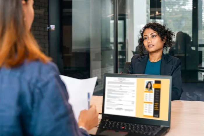 A professional woman is interviewed in an office setting; interviewer holds a resume, visible on laptop